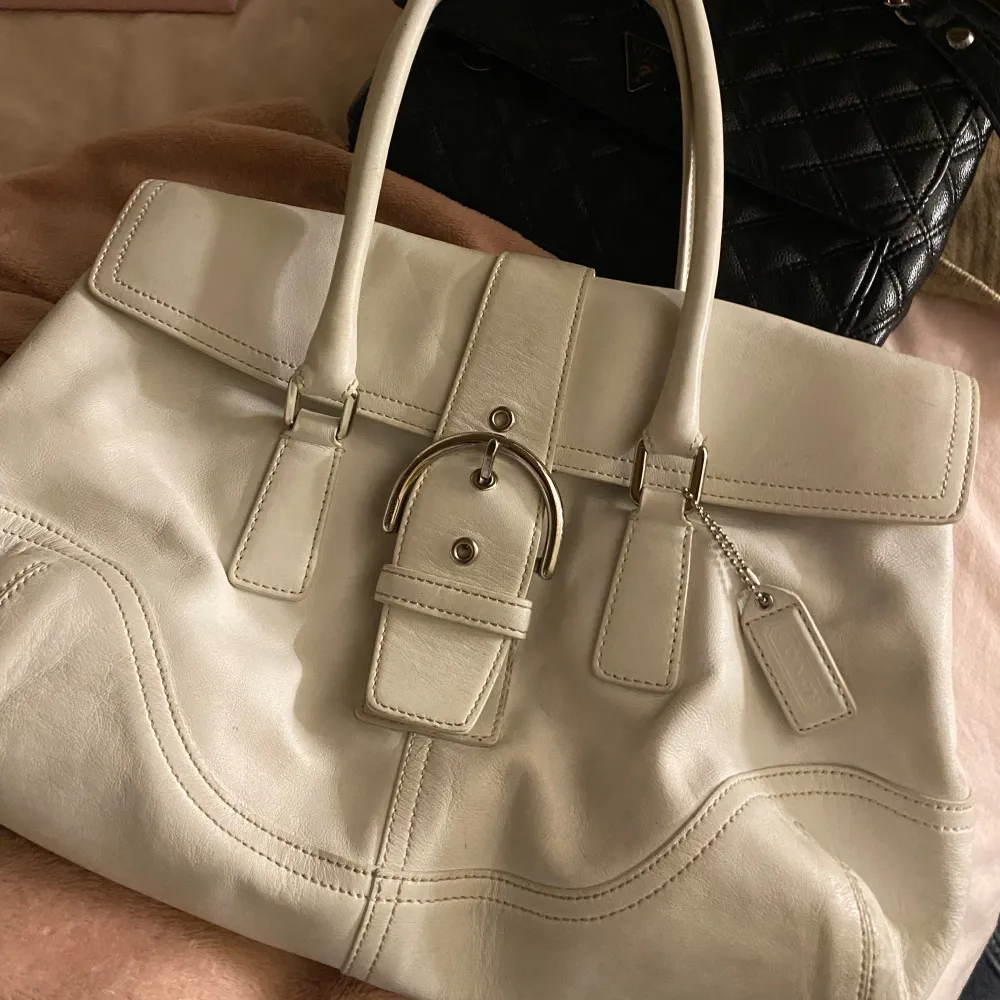 Säljer en elegant vit handväska från Coach i skinn. Väskan har en stilren design med ett dekorativt bälte och en praktisk dragkedja inuti. Perfekt för både vardag och fest! 👜✨. Väskor.