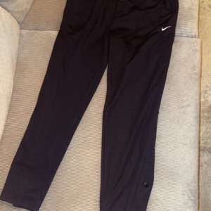 Svarta träningsbyxor från Nike - Säljer ett par svarta träningsbyxor från Nike i Dri-FIT material. Byxorna har en normal passform med knappar längs sidorna för extra ventilation. Perfekta för träning eller en avslappnad dag. De är helt nya med taggar kvar!