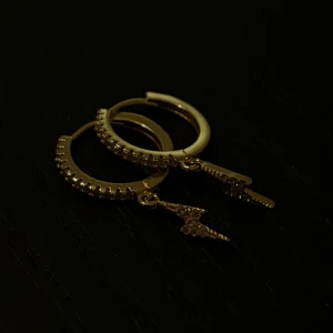 Guldörhängen med blixtberlocker - Snygga guldörhängen med små blixtformade berlocker. De har en glittrig yta som ger en extra touch av stil. Perfekta för att liva upp vilken outfit som helst, oavsett om det är till vardags eller fest. Ett måste för alla som älskar unika smycken! 