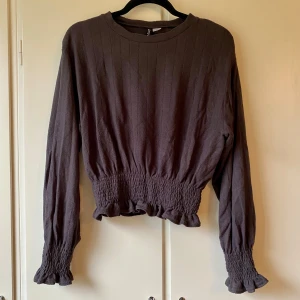 Grå långärmad tröja med volangdetaljer - Säljer en fin grå tröja med långa ärmar och volangdetaljer vid midjan och ärmsluten. Den har en ribbad struktur och en elastisk midja som ger en snygg passform. 