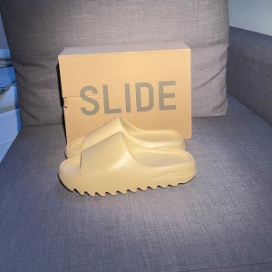Beige Yeezy Slide från Adidas - Säljer ett par snygga och bekväma Yeezy Slide i beige från Adidas. Perfekta för sommaren med sin stilrena design och mjuka material. De har en modern look med öppen tå och räfflad sula för extra grepp. Kommer i originalförpackning.