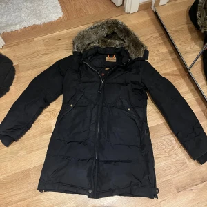 Svart vinterjacka från Parajumpers - Säljer en svart Parajumpers vinterjacka i mycket bra skick. Jackan har en mysig pälsfodrad huva och praktiska fickor med knappar. Perfekt för kalla vinterdagar! Till jackan kommer även en päls krage som jag för tillfället inte kunde hitta, men vid intresse kan jag försöka få tag på den. Annars kan man lägga till sin egna pälskräge ifall man har och vill. Jacken är helt äkta och köptes i butik för ungefär 6000