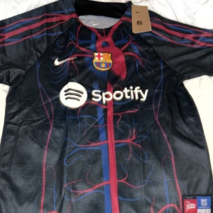 Svart och röd FC Barcelona fotbollströja - Säljer en ny FC Barcelona tröja i samarbete med Patta. Den är svart med röda och blå detaljer och har klubbens emblem och Spotify-logga på framsidan. Tröjan är kortärmad och har en modern design med Dri-FIT material för bästa komfort. Perfekt för alla Barça-fans! Helt ny! True to size