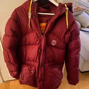 Vinröd dunjacka från Fjällräven - Säljer en supervarm och snygg vinröd dunjacka från Fjällräven. Perfekt för kalla vinterdagar! Jackan har en praktisk huva och gula dragskor för justering. Den är i mycket bra skick och har både dragkedja och knappar framtill. Passar perfekt för vinterns alla äventyr! ❄️