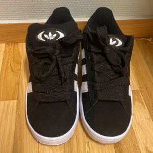 Svarta Adidas sneakers - Säljer ett par snygga svarta Adidas sneakers i mycket bra skick. De har de klassiska tre vita ränderna på sidan och en vit sula. Perfekta för vardagsbruk och ger en sportig look. Skorna har snörning och är bekväma att gå i. Passar till både jeans och shorts!