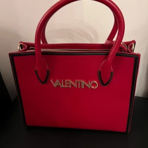 Röd handväska från Valentino - Säljer en snygg röd handväska från Valentino som är helt ny. Väskan har ett stilrent och elegant utseende med guldiga detaljer och en praktisk dragkedja upptill. Perfekt för både vardag och fest!