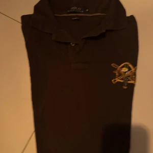 Brun pikétröja från Ralph Lauren - Säljer en snygg brun pikétröja från Ralph Lauren. Den har en klassisk krage och ett broderat emblem på bröstet. Perfekt för en stilren och avslappnad look. Tröjan är i bra skick och passar perfekt för både vardag och lite finare tillfällen.