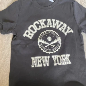 Svart t-shirt med Rockaway New York-tryck från Divided - Säljer en svart t-shirt från Divided med ett coolt Rockaway New York-tryck i vitt. Perfekt för en avslappnad stil och passar till både jeans och shorts. T-shirten är i bomull och har en normal passform. Perfekt för vardagsbruk!