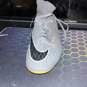 Gråa Nike fotbollsskor - Säljer ett par gråa Nike fotbollsskor i nyskick. De har en snygg svart och orange detalj på sidan och är perfekta för fotbollsträning. Skorna har snörning och en tight passform som ger bra stöd på planen. Sulan är designad för optimalt grepp på gräs.