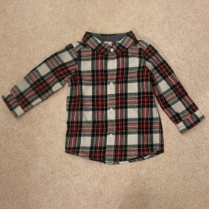 Rutig skjorta från H&M - Säljer en superfin rutig skjorta från H&M i storlek 80, perfekt för små äventyrare! Skjortan har ett klassiskt rutmönster i rött, grönt och vitt med knappar framtill. Den är långärmad och gjord i mjukt bomullsmaterial, vilket gör den både bekväm varm