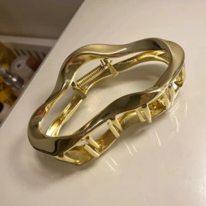 Guldig hårklämma - Snygg och stilren guldig hårklämma i metall med vågig design. Perfekt för att hålla håret på plats med stil, oavsett om det är till vardags eller fest. Klämman har en glansig finish som ger en lyxig känsla. Ett måste för alla som älskar att piffa upp sin look med accessoarer! Aldrig använd