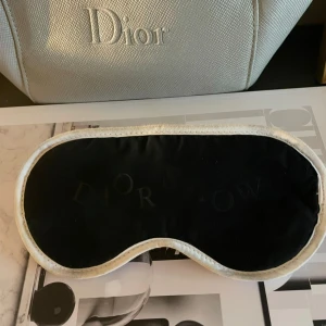 Sovmask från Dior - Elegant sovmask från Dior i svart och beige. Masken har en mjuk och bekväm passform med Dior-logotypen snyggt placerad. Perfekt för en lyxig och avkopplande sömnupplevelse.