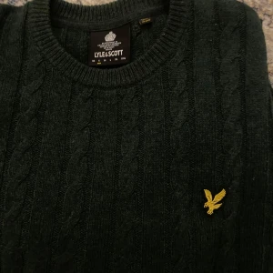 Mörkgrön stickad tröja från Lyle & Scott - Säljer en mörkgrön stickad tröja från Lyle & Scott med kabelstickat mönster och ett gult broderat emblem på bröstet. Tröjan har långa ärmar och ribbstickade muddar vid ärmslut och nederkant. Fräsch färg!