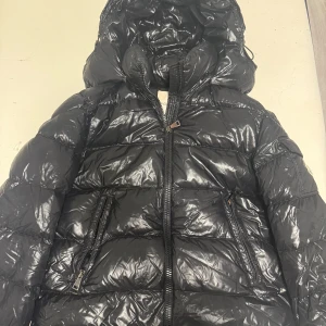 Svart dunjacka från Moncler (Maire) - Snygg och glansig svart dunjacka från Moncler med huva och dragkedja. Jackan har praktiska fickor med dragkedjor och en logotyp på ärmen. Perfekt för kyliga dagar. Säljs idag på Monclers Hemsida för ca. 18 800kr