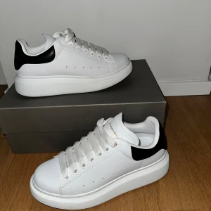 Vita sneakers från Alexander McQueen - Snygga vita sneakers från Alexander McQueen med svart detalj på hälen. Skorna har en klassisk design med snörning och en tjock sula för extra komfort. Perfekta för en stilren look. Köpt för 5300kr.  Sko lådan är tyvärr borttappat. Fick som present och är knappast använd som man ser på bilden. Kan sälja den billigare vid snabb affär.