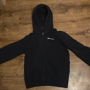 Svart hoodie från Champion - Säljer en svart hoodie från Champion med dragkedja och huva. Den har långa ärmar och Champion-loggan broderad på bröstet och ärmen. Perfekt för en avslappnad stil.