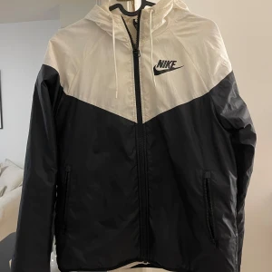 Svart och vit vindjacka från Nike - Snygg svart och vit vindjacka från Nike med dragkedja och huva. Jackan har en klassisk design med Nike-logga på bröstet och praktiska fickor med dragkedja. Perfekt för blåsiga dagar.