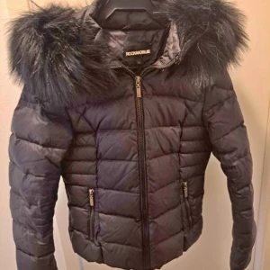 Svart dunjacka från Rockandblue, strl: 36/34 - Snygg svart dunjacka från Rockandblue med pälskantad huva. Jackan har dragkedja framtill och två fickor med dragkedjor. Perfekt för kyliga dagar med sin quiltade design och stilrena look. Den är strl 36 men sitter mer som en 34.
