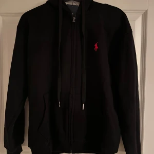 Svart hoodie från Ralph Lauren - Snygg svart hoodie från Ralph Lauren med dragkedja och röd logga på bröstet lappen sitter kvar på. Säljer för att den var för liten för mig.