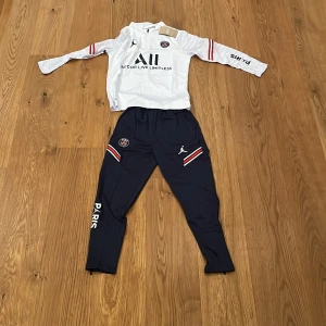 Vit/blå PSG Tracksuit  - Snygg vit långärmad PSG tröja med Jordan-logga och texten 'Accor Live Limitless' på framsidan. Röd och svart randdetalj på ärmarna och 'Visit Rwanda' på baksidan. Perfekt för fans av Paris Saint-Germain. Tracksuiten  är helt ny och passar från 135-160 (cm) Har ni några funderingar så ska ni inte tveka på att fråga! 