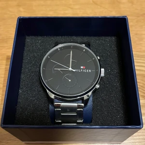 Tommy Hilfiger Klocka - Säljer snygg klocka från Tommy Hilfiger med svart urtavla och silverfärgat metallarmband. Mycket bra skick, kommer i originallåda med segment för storleksjusteringar. OBS batteriet är slut och måste bytas (därför är den billigare)