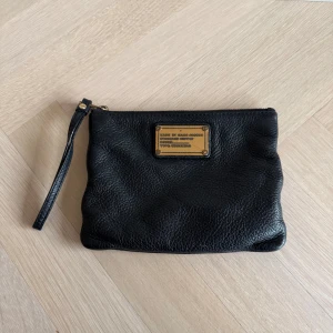 Svart clutch från Marc by Marc Jacobs - Snygg svart väska i skinn från Marc by Marc Jacobs med dragkedja och ett stilrent märke på framsidan. Insidan har ett coolt mönster med bokstäver. Perfekt för att hålla ordning på dina saker med stil. 20 x 15 cm.