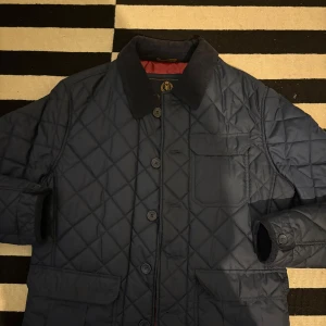 Blå quiltad jacka från Henri Lloyd - Säljer en snygg blå quiltad jacka från Henri Lloyd. Jackan har en klassisk krage och knappar framtill. Den är fodrad med rött tyg och har praktiska fickor. Perfekt för kyligare dagar! Inte använd så mycket men kör på en annan jacka iår istället o vill ha pengarna. Skriv om ni har frågor!