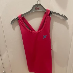 Rosa linnetopp från Li-Ning - Säljer en sportig rosa linnetopp från Li-Ning med ett blått logotryck på bröstet. Perfekt för träning med sin bekväma passform och ärmlösa design.