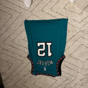 Turkos basketlinne från NBA - Säljer ett turkost basketlinne med nummer 12 och namnet Morant. Linnet har svarta och röda detaljer längs ärmhålen och nederkanten. Perfekt för basketfans som vill visa sitt stöd.