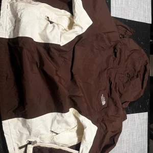 Brun och vit jacka från The North Face - Säljer en brun och vit skaljacka från The North Face. Jackan har dragkedja och flera praktiska fickor. Perfekt för utomhusaktiviteter med sin funktionella design. Säljs på grund av att tidigare ägare sålde den som en jacka för män när den egentligen var för kvinnor.