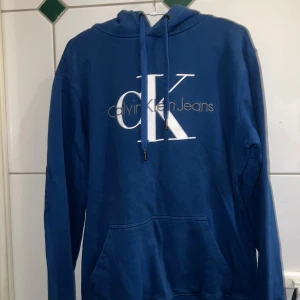 Blå hoodie från Calvin Klein Jeans - Snygg blå hoodie från Calvin Klein Jeans med stor logga på framsidan. Tröjan har en justerbar huva med dragsko och långa ärmar. Perfekt för en avslappnad stil.