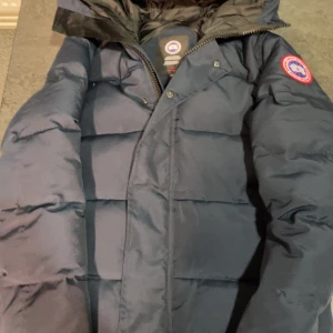 Mörkblå dunjacka från Canada Goose - Säljer en mörkblå dunjacka från Canada Goose med lång ärm och dragkedja. Jackan har en broderad logotyp på ärmen och är perfekt för kalla vinterdagar.