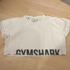 Vit croppad t-shirt från Gymshark - Säljer en vit croppad t-shirt från Gymshark i storlek XS. T-shirten har korta ärmar och ett stort tryck med märkesnamnet längst ner. Perfekt för en avslappnad stil eller träning.