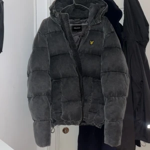 Grå pufferjacka från Lyle & Scott - Säljer en grå pufferjacka från Lyle & Scott med huva och dragkedja. Jackan har en broderad logotyp på bröstet och är tillverkad i 100% polyester. Perfekt för kyligare dagar. Köpt för 1699kr
