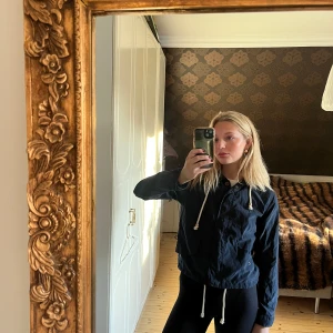 marinblå vindjacka med dragkedja - Snygg marinblå vindjacka med dragkedja och justerbar dragsko i midjan. Perfekt för blåsiga dagar och har en stilren design som passar till det mesta. Jackan har långa ärmar och en bekväm passform.