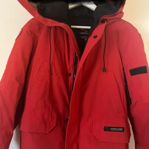 Röd dunjacka från Canada Goose - Säljer en röd dunjacka från Canada Goose med huva och pälsdetaljer. Jackan har dragkedja och knappar framtill samt flera praktiska fickor. Perfekt för kalla vinterdagar.