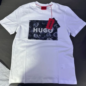 Hugo t-shirt - Helt ny Hugo T-shirt. Snygg  vit t-shirt från Hugo med ett stort blommigt tryck i svart och grått på framsidan. Perfekt för en stilren och modern look. T-shirten  har en klassisk passform. Köpt för 789kr