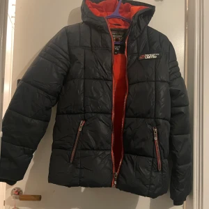 Svart dunjacka från Authentic Outdoor Co. - Säljer en svart dunjacka från Authentic Outdoor Co. med orange foder och dragkedja. Jackan har en quiltad design och huva, perfekt för kyliga dagar. Två praktiska fickor med dragkedja framtill.