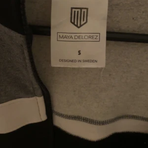Svart och grå hoodie från Maya Delorez - Snygg svart och grå hoodie från Maya Delorez med dragkedja och kontrasterande vita detaljer. Perfekt för en avslappnad stil. Designad i Sverige.