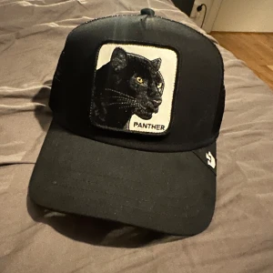 Svart keps med pantermotiv från The farmFarm - Snygg svart keps med ett broderat pantermotiv på framsidan. Kepsen har justerbar passform med nätmaterial baktill för extra ventilation. Perfekt för en cool och avslappnad stil.