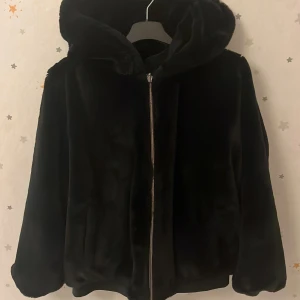Svart pälsjacka med dragkedja - Snygg svart pälsjacka med huva och dragkedja framtill. Jackan har en mjuk och lyxig känsla, perfekt för kyligare dagar. Den har en stilren design med långa ärmar och en bekväm passform. Det är liksom 2 jackor i en🤩