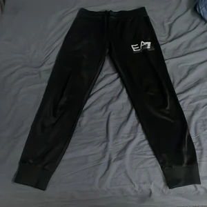 Svarta mjukisbyxor från Emporio Armani - Snygga svarta mjukisbyxor från Emporio Armani med EA7-logga på benet. Byxorna har en bekväm passform med elastisk midja och muddar vid bensluten. Perfekta för en avslappnad stil.