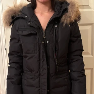 Svart dunjacka från Hollies - Säljer en svart dunjacka från Hollies med en stor, fluffig päls på huvan. Jackan har dragkedja och knappar framtill samt flera praktiska fickor. Perfekt för kalla vinterdagar.