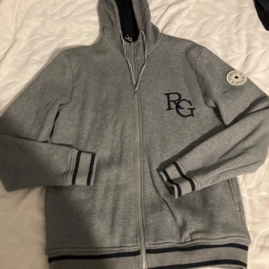 Grå hoodie med dragkedja från Redgreen - Säljer en stilren grå hoodie från Redgreen med dragkedja och broderad logga på bröstet. Tröjan har långa ärmar och en rund patch på ärmen. Perfekt för en avslappnad stil.