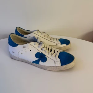 Vita och blå sneakers med klöverdetalj - Aldrig använda, helt nya. Har två likadana par, därav två annonser. Storlek 42. Box finns ej men skickas med låda.
