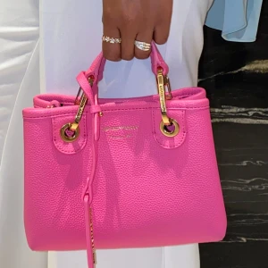 Rosa Armani handväska med gulddetaljer - Snygg rosa handväska med gulddetaljer och dragkedja. Perfekt för att ge en färgklick till din outfit. Den har en stilren design med praktiska handtag.