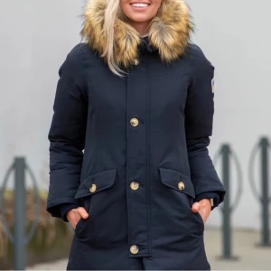 Svart dunjacka från Svea - Säljer en snygg svart dunjacka från Svea med pälskantad huva och knappar framtill. Jackan har långa ärmar och praktiska fickor med lock. Perfekt för kalla dagar!