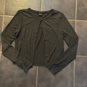 Mörkgrå kofta från Gina Tricot - Säljer en stilren svart kofta från Gina Tricot. Koftan har långa ärmar och en enkel knapp framtill. Perfekt för lager-på-lager och passar till många olika outfits.