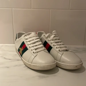 Gucci ace skor - Snygga vita sneakers med gröna och röda ränder samt en guldfärgad bi-detalj på sidan. Skorna har snörning och en kontrasterande häl i svart och rött. Perfekta för en stilren look. Gucci ace skor säljs för 2199 kan tänka mig gå för 1999 storlek 41 men passar 42