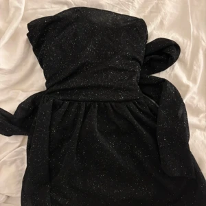 Svart glittrig klänning - Elegant svart klänning med glittrande detaljer. Den har en knytning framtill och är perfekt för festliga tillfällen. Klänningen är axelbandslös och har en vacker drapering.